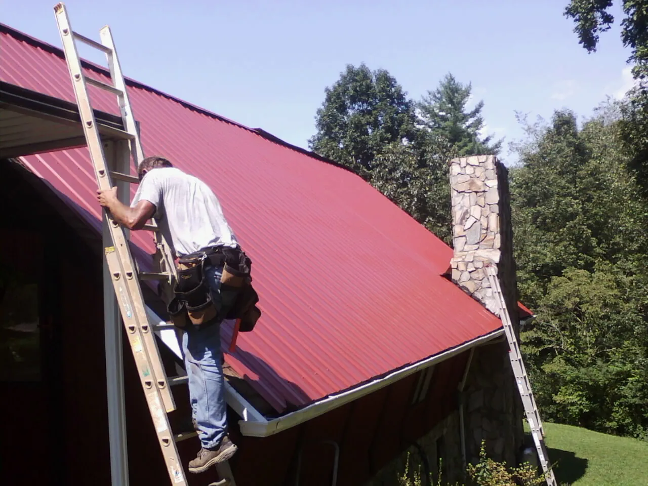 Roofer on a ladder beside a red metal roof and stone chimney — Soffit & Fascia Repair by Roofing Brothers in Ellston