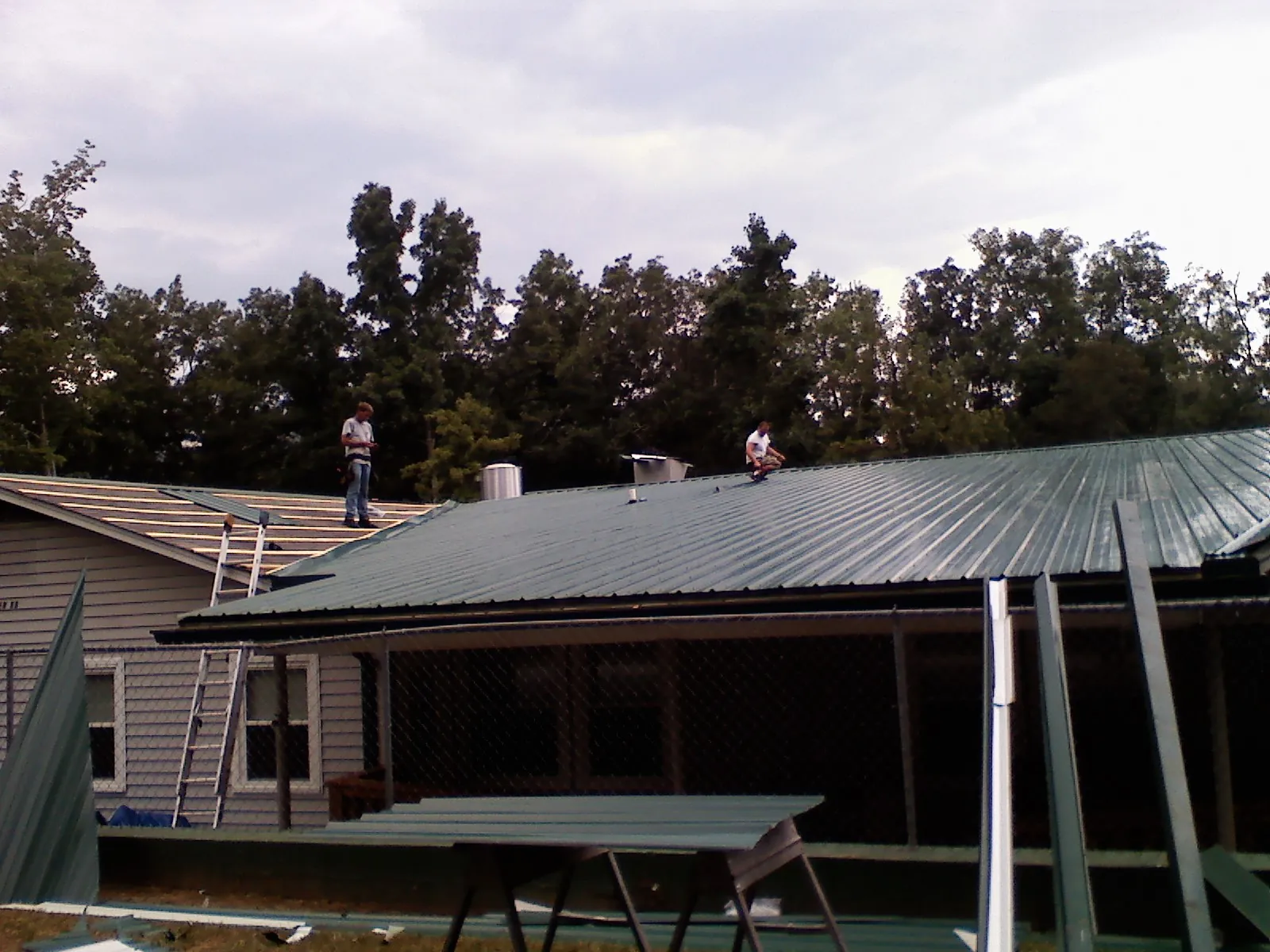 Two roofers installing green metal panels above a covered porch in Ellston